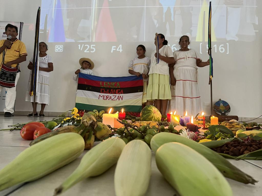 En primer plano, altar formado por los pueblos indígenas con mazorcas de maíz, flores, frutas y velas. Al fondo, mujeres sosteniendo banderines y una joven y un niño con una bandera que dice Kakawira, Morazán.