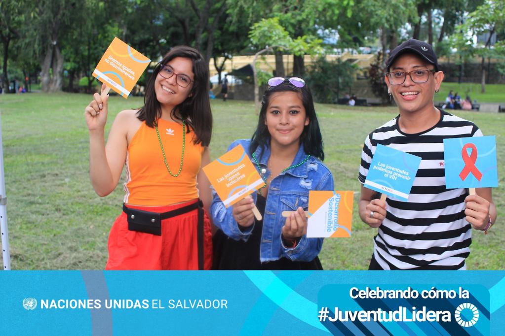 En primer plano dos jóvenes mujeres y un joven hombre sostienen rótulos sobre igualdad, liderazgo juvenil y prenvención del VIH. Al fondo el se ven árboles y en la parte baja de la fotografía está el logotipo de las Naciones Unidas y la frase "Celebrando cómo la juventud lidera"