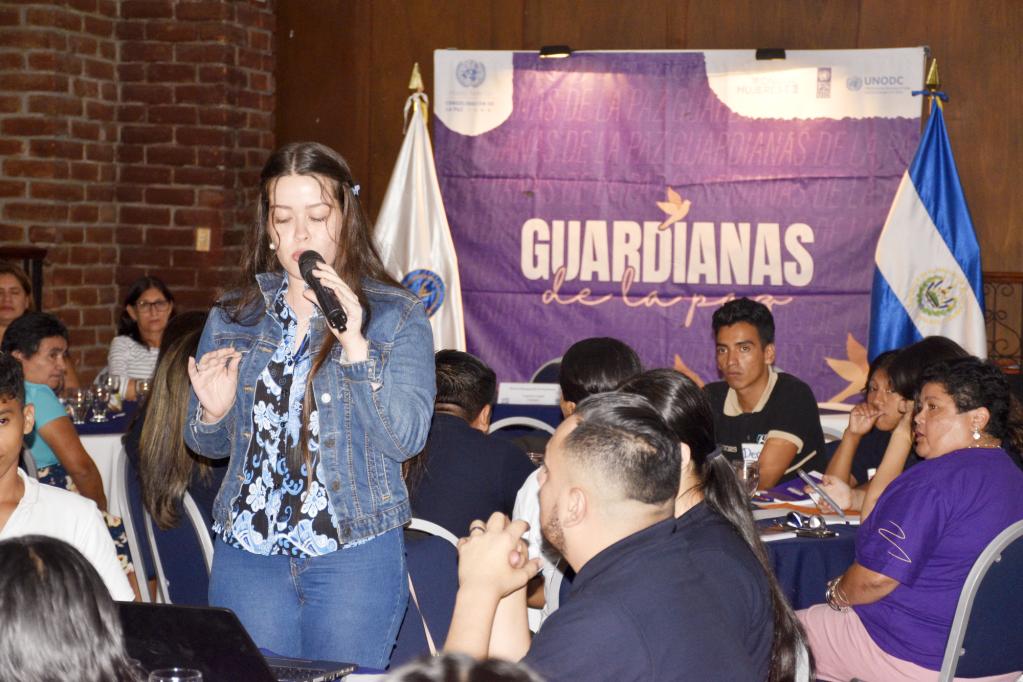 Mujer joven en primer plano con un micrófono expone los resultados de su mesa de trabajo. Grupo de personas sentadas en mesas redondas a su alrededor la observan. Al fondo un banner en el que se lee Guardianas de la paz.