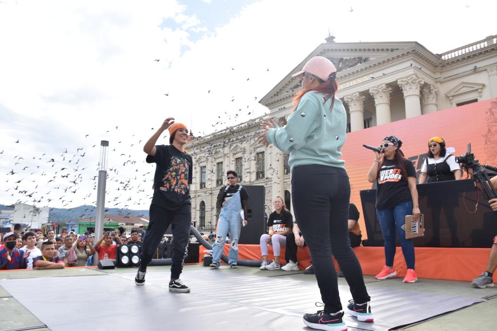 Dos jóvenes sobre un escenario colocado frente al Palacio Nacional se acercan para darse un abrazo luego de finalizar un duelo de break dance. Al fondo, las juezas del duelo, una pantalla led con color naranja y el Palacio Nacional.