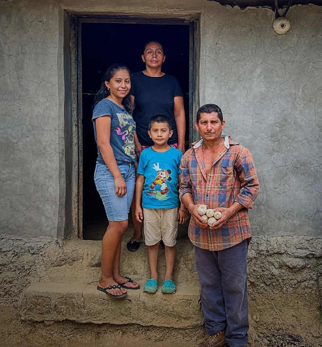 Una familia posa en la puerta de su casa de adobe. El padre sostiene unas mazorcas de maíz.