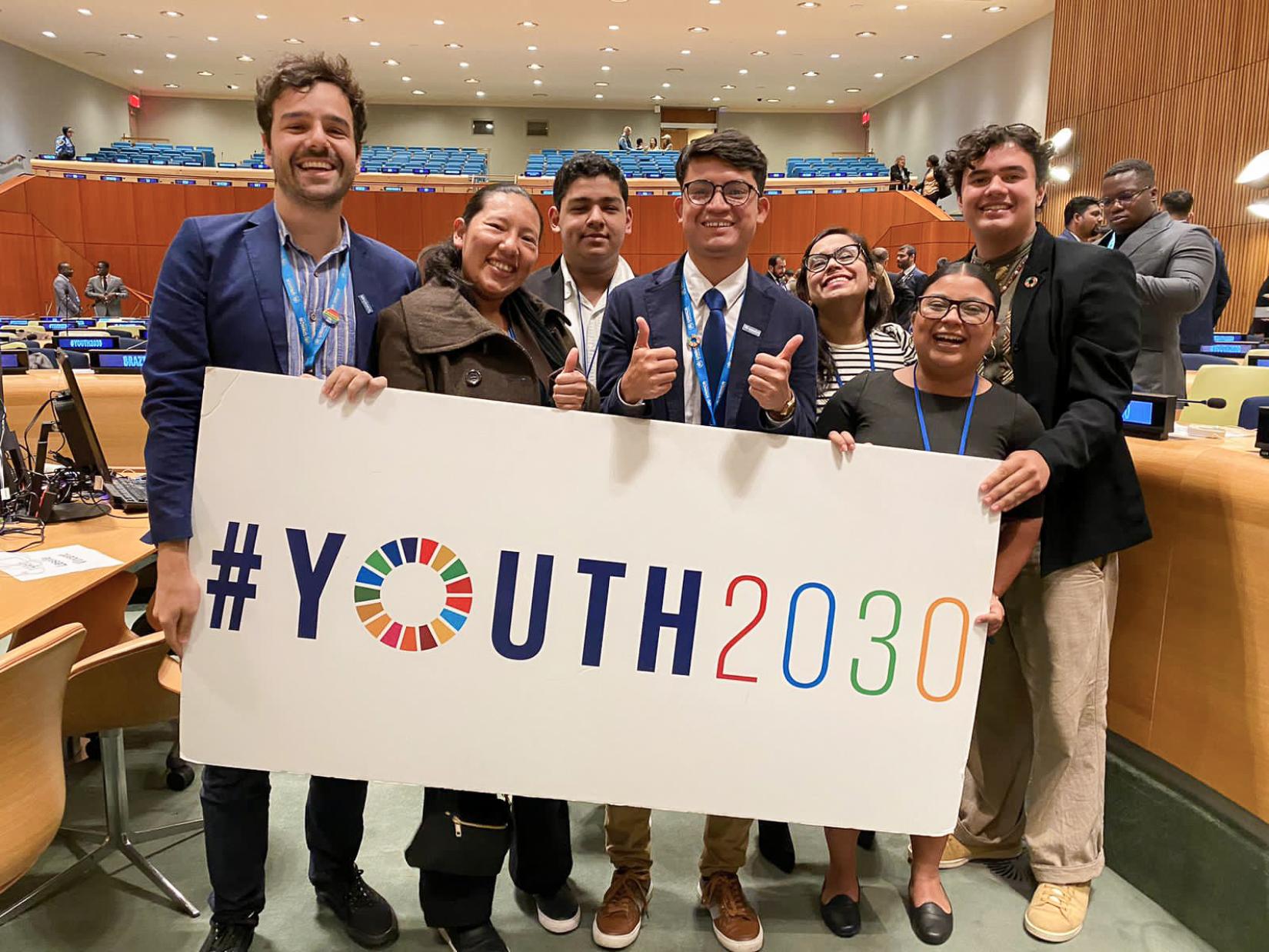 En primer plano, un grupo de jóvenes sostiene un cartel con la etiqueta #Youth2030 (#Juventud2030), al fondo el salón donde se desarrolló la Cumbre de la Juventud en la sede de las Naciones Unidas en Nueva York.