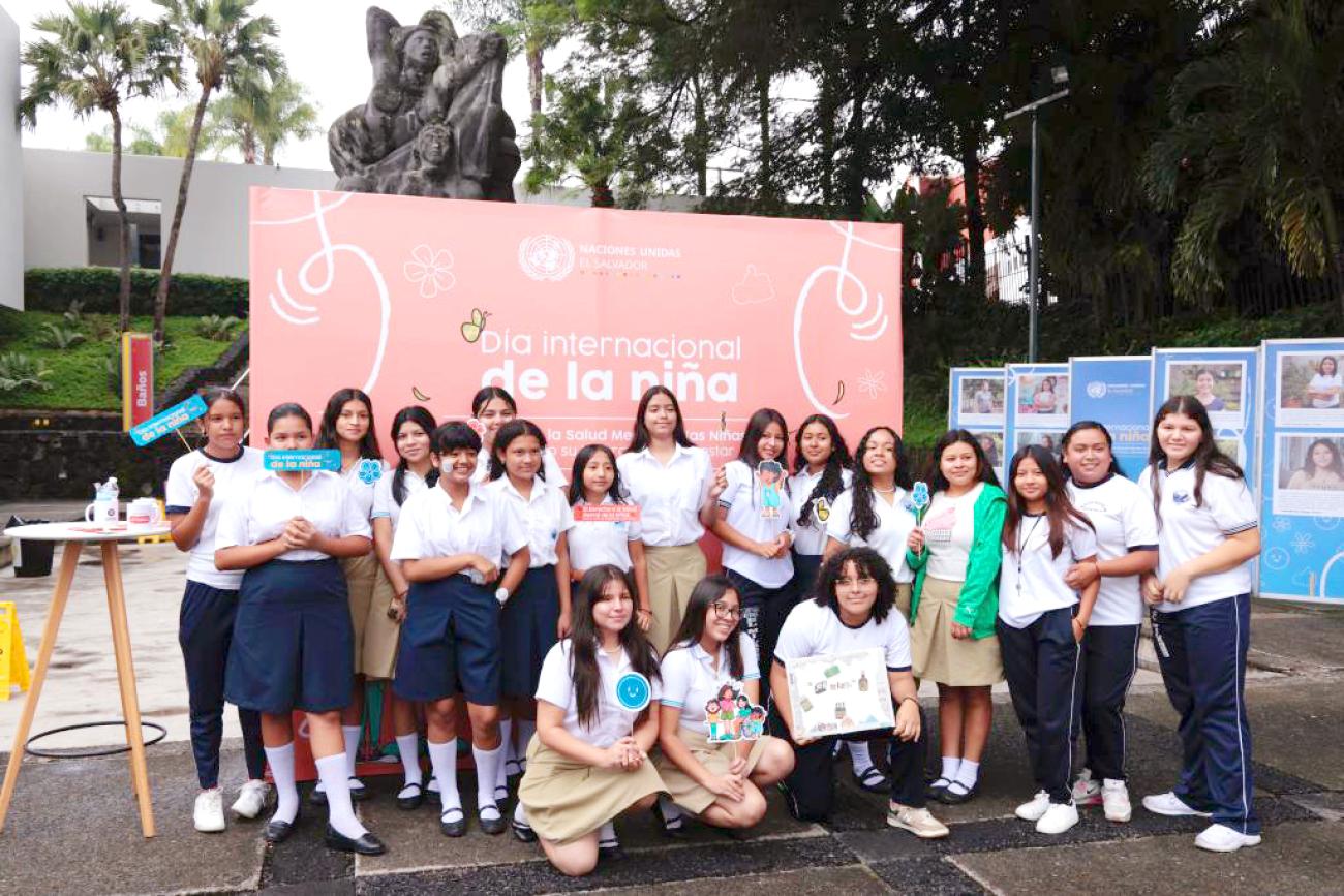 Grupo de niñas posa para la fotografía con rótulos sobre el día de la niña, al fondo un banner que dice Día internacional y nacional de la niña y una exposición con fotografías de otras niñas y testimonios.