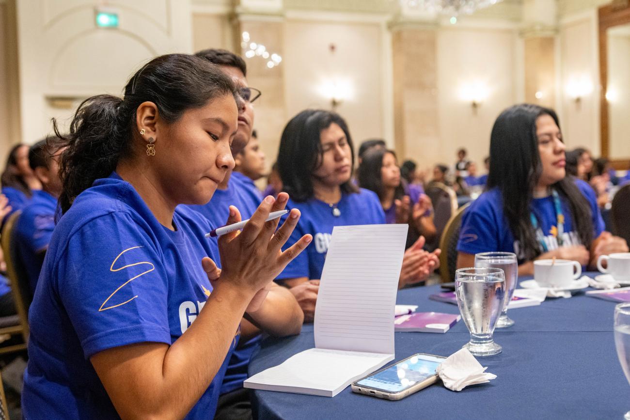 En primer plano, aparece una joven tomando nota en su mesa, con otras jóvenes al fondo.