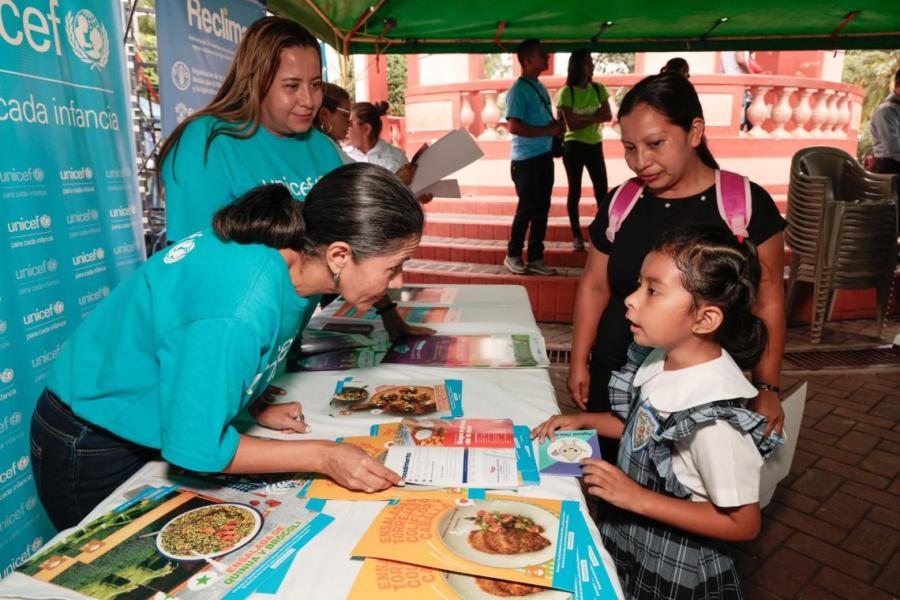 Una niña con su madre recibe un recetario de alimentación saludable de dos oficiales mujeres de UNICEF en un stand ubicado en un parque público.
