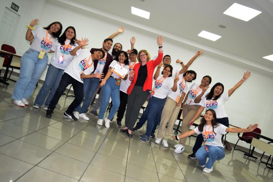 Foto de un grupo de jóvenes, mujeres y hombres, salundando alegres a la cámara, en un salón y con la representante del UNFPA al centro.