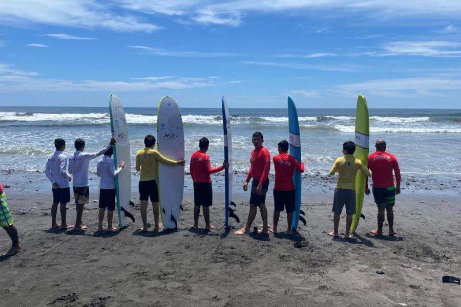 Un grupo de jóvenes con tablas de Surf están a la orilla del mar, observando hacia la playa antes de comenzar a poner en práctica lo aprendido en las escuelas de Surf