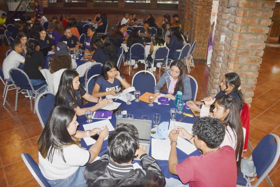 Ocho jóvenes sentados en una mesa redonda diálogando, al fondo se aprecian otras tres mesas redondas con otros grupos de jóvenes y víctimas intercambiando experiencias.