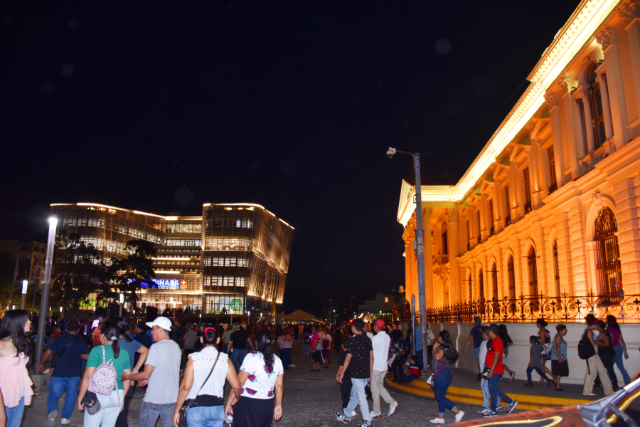 En primer plano, el Palacio Nacional ilimunado de naranja, personas caminando alrededor y al fondo la Bilioteca Nacional de El Salvador.