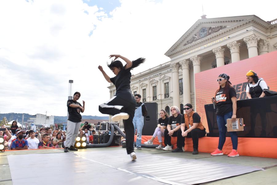 En primer plano, una joven bailando breack dance sobre una tarima, enfrente de tres juezas, la DJ y su competidora, al fondo el Palacio nacional.
