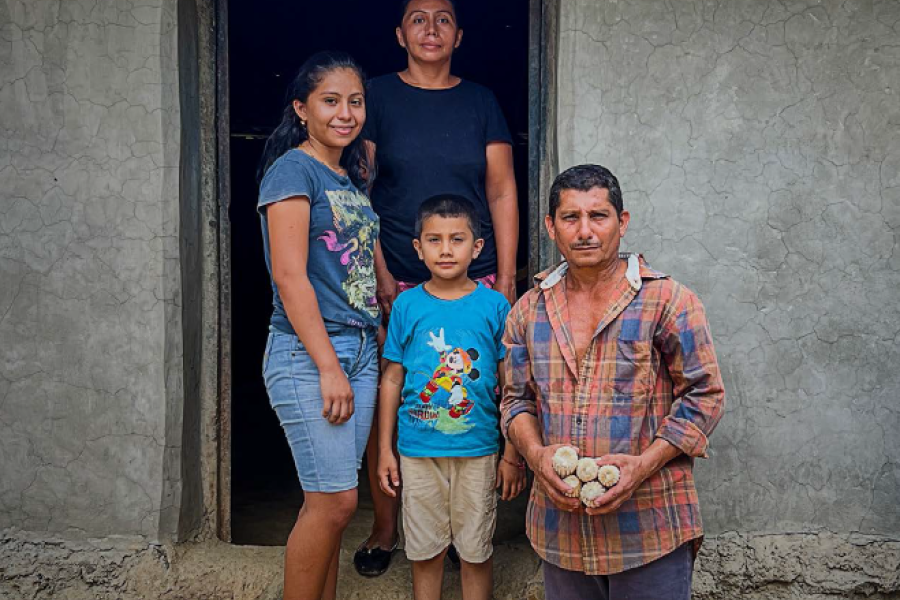 Una familia posa en la puerta de su casa de adobe. El padre sostiene unas mazorcas de maíz.