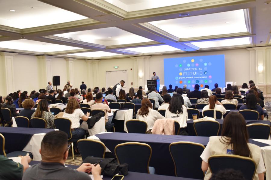 Foto panorámica de asistentes al evento Imaginar el futuro. Al fondo, el Coordinador Residente en el podio ubicado en la tarima, expone su mensaje a las personas asistentes al evento.