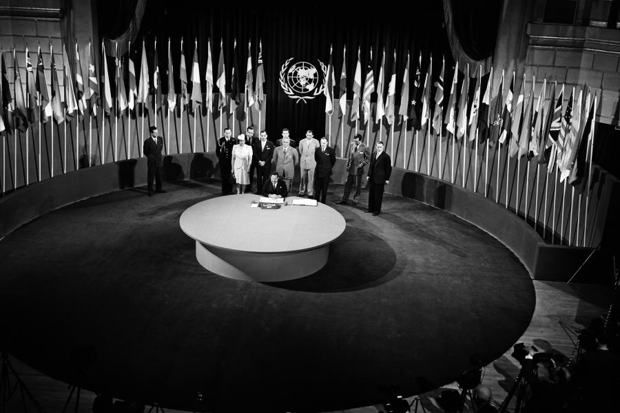 Fotografía en blanco y negro del momento en que se firmó la Carta de las Naciones Unidas. Los firmantes alrededor de una mesa redonda y rodeados por banderas de los países que integraban entonces las Naciones Unidas