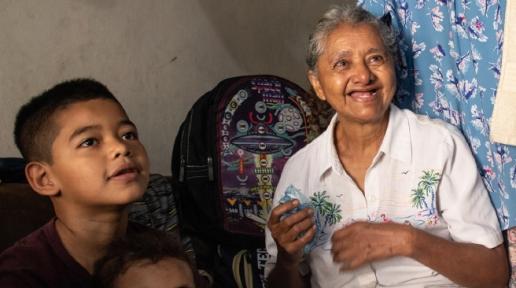 Texto que dice Plan de Respuesta Humanitaria El Salvador. Ciclo del programa humanitario 2023. Abajo, fotografía de un niño, una niña y una mujer adulta mayor sentados y sonriendo y viendo hacia otra persona que está con ellos fuera del encuadre de la foto.