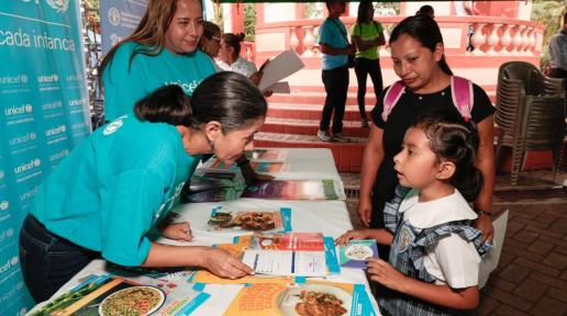 Una niña con su madre recibe un recetario de alimentación saludable de dos oficiales mujeres de UNICEF en un stand ubicado en un parque público.