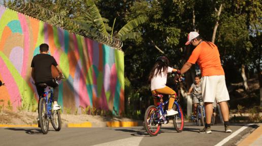 Un niño y una niña ayudada por su padre manejan una bicicleta en la calle con un mural de distintos colores al fondo.