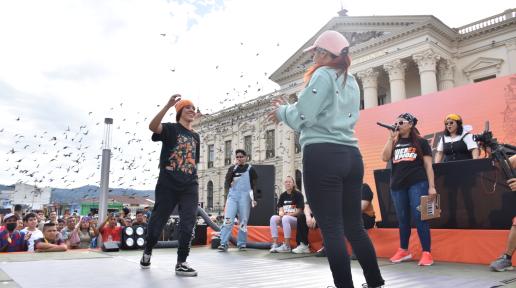 Dos jóvenes sobre un escenario colocado frente al Palacio Nacional se acercan para darse un abrazo luego de finalizar un duelo de break dance. Al fondo, las juezas del duelo, una pantalla led con color naranja y el Palacio Nacional.